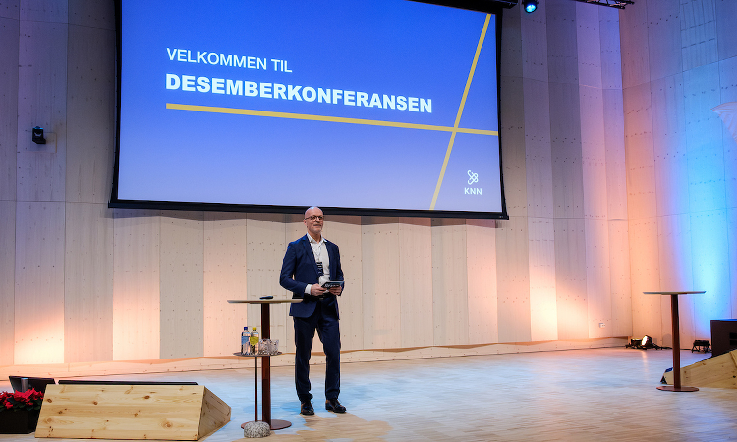 Mann i mørk blå dress ser utover et rom og smiler. Han står på et scnenegulv. Bak ham står det 