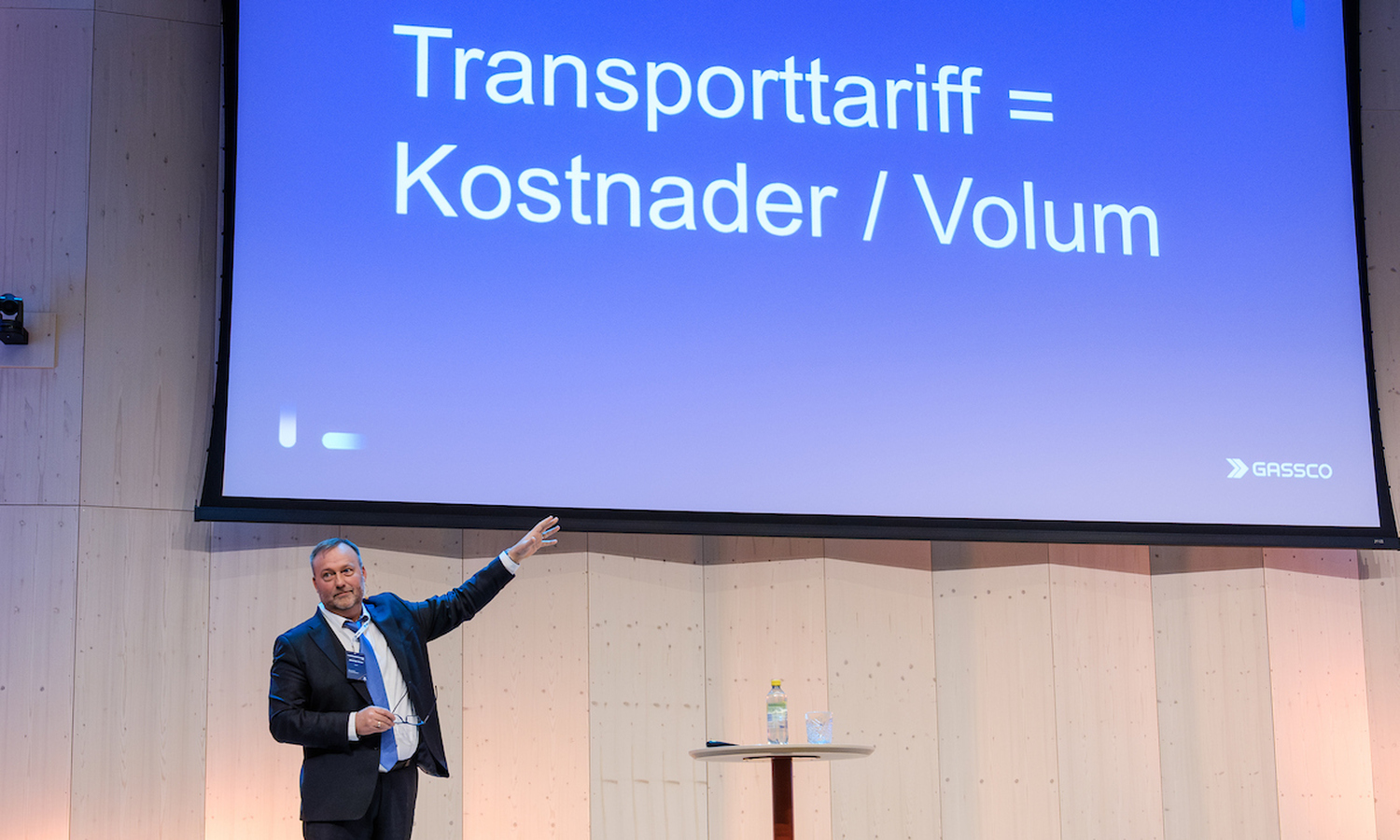 mann i dress peker mot et blått lerret med hvit påskrift der det står: Transporttariff = kostnader / Volum