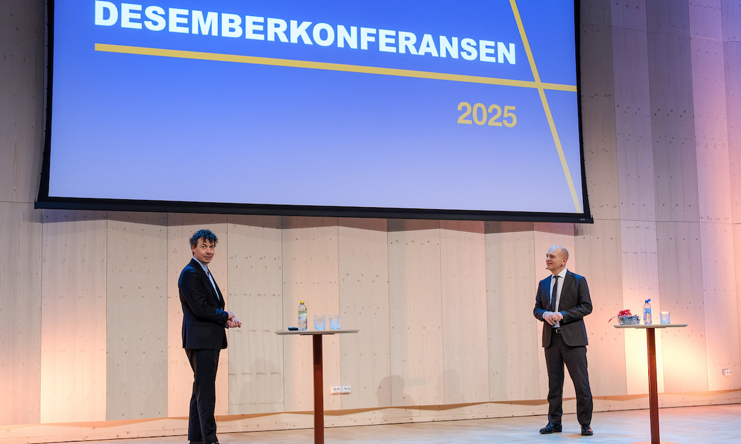 To menn i mørke dresser på en scene av heltre, omgitt av vegger i heltre og et lerret med påskriften 