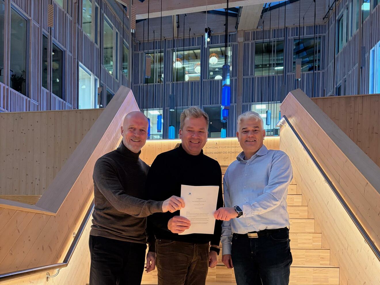 tre men står i en trapp laget av massivt tre og holder en bunke papirer sammen. 