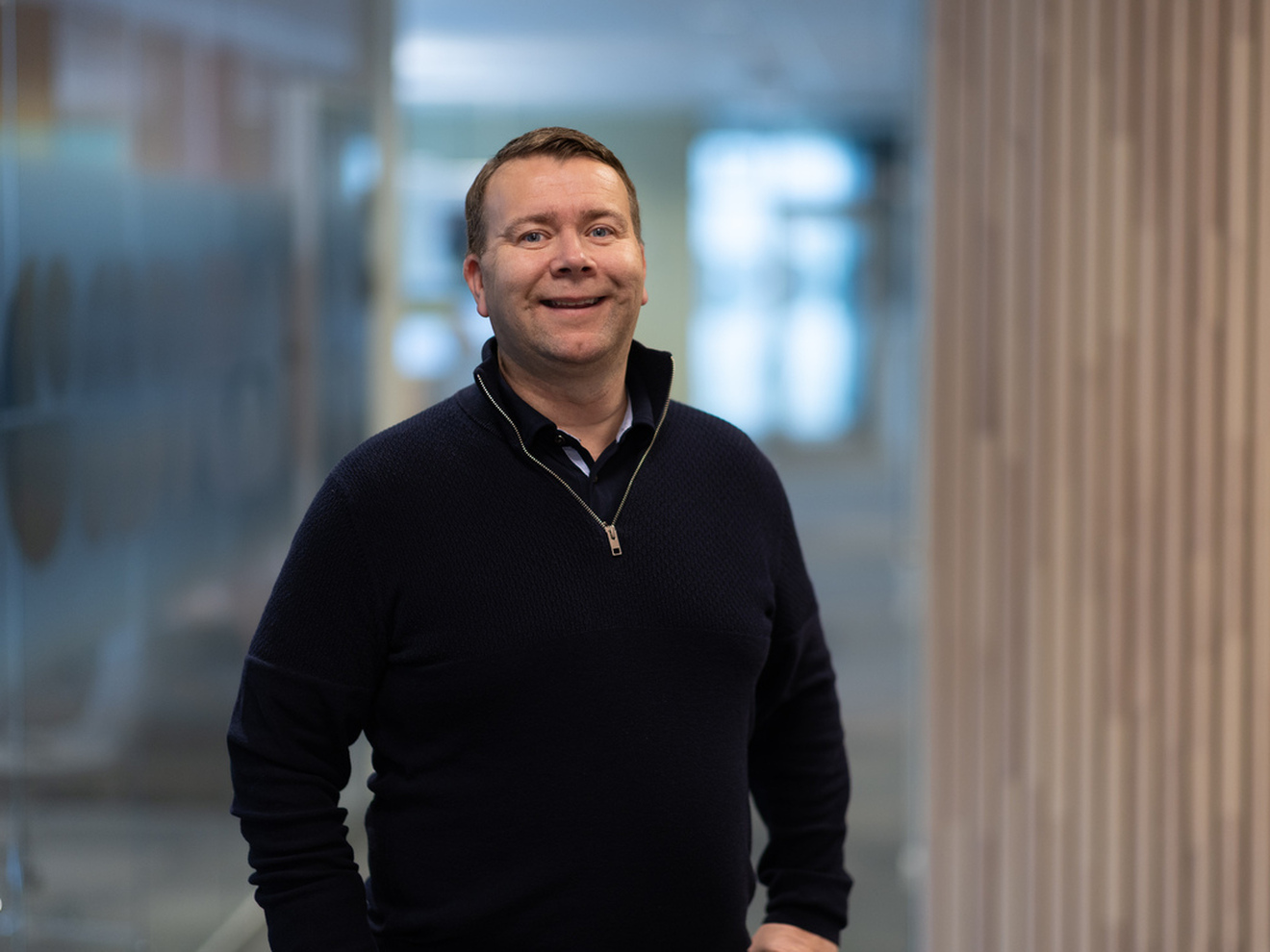 en mann i sort genser smiler mot kamera. Bakgrunnen er uskarp og ser ut til å være i en korridor