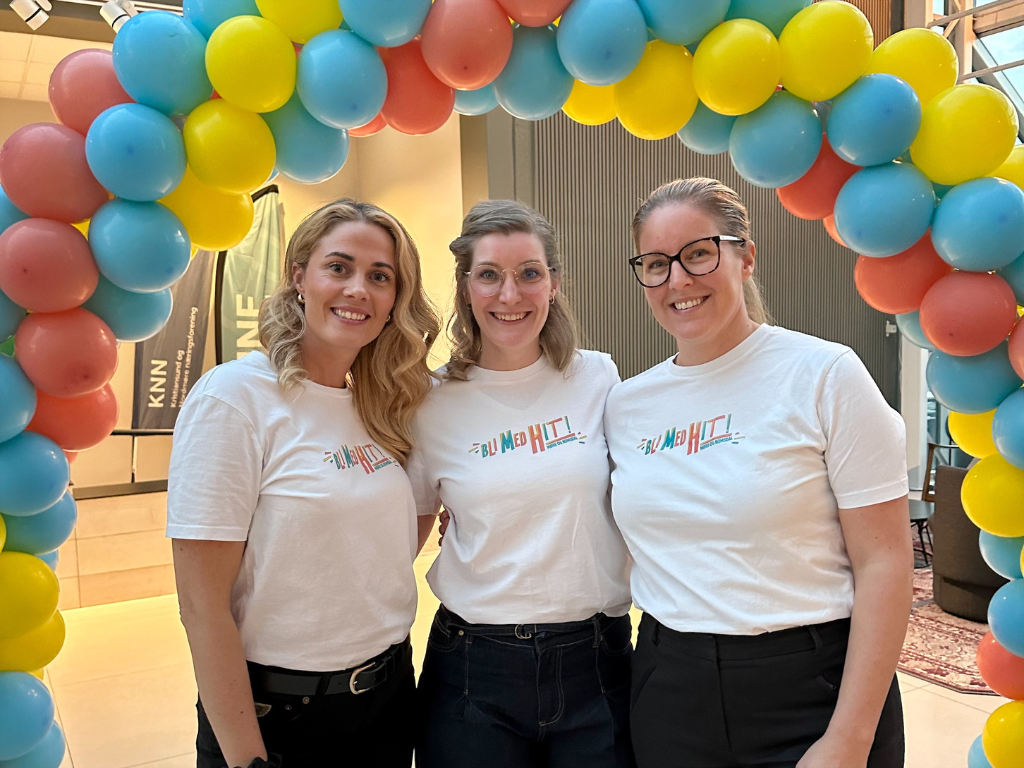 Tre kvinner i hvite t-.skjorter med påskriften BLI MED HIT, står under en ballongbue bestående av gule, røde og blå ballonger, smiler mot kamera. 
